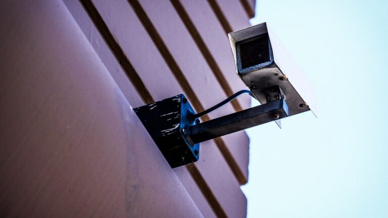 Câmera de vigilância presa à fachada de um prédio, vista de baixo, com parede lilás, imagem ilustrativa do artigo sobre Prevencao a lavagem de dinheiro na empresa: deveres da Lei